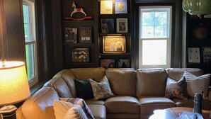 Un salon chaleureux avec un canapé d’angle en cuir, une petite table d’appoint, une lampe et une bibliothèque contenant des photos encadrées et des livres.