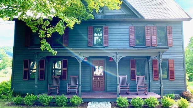 Une maison à deux étages avec un porche, de nombreuses fenêtres dotées de volets et une porte d’entrée.