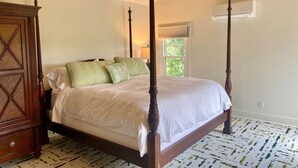 Un lit à baldaquin avec une structure en bois, une literie blanche et des oreillers verts. Une table de chevet en bois avec une lampe se trouve à côté du lit. La pièce a un plafond en pente avec des poutres apparentes et une fenêtre avec des stores.
