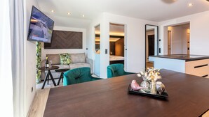 Una sala moderna con un sofá, una mesa de comedor con objetos decorativos, un televisor montado en la pared y una cocina visible a través de una puerta abierta.