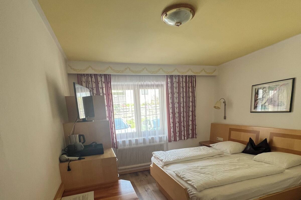 Habitación de hotel con escritorio de madera, cama, silla, ventana con cortinas y una lámpara en el techo.