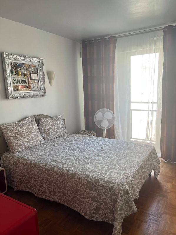 A bedroom with a bed, a red chair, a wall-mounted fan, and a window with curtains.