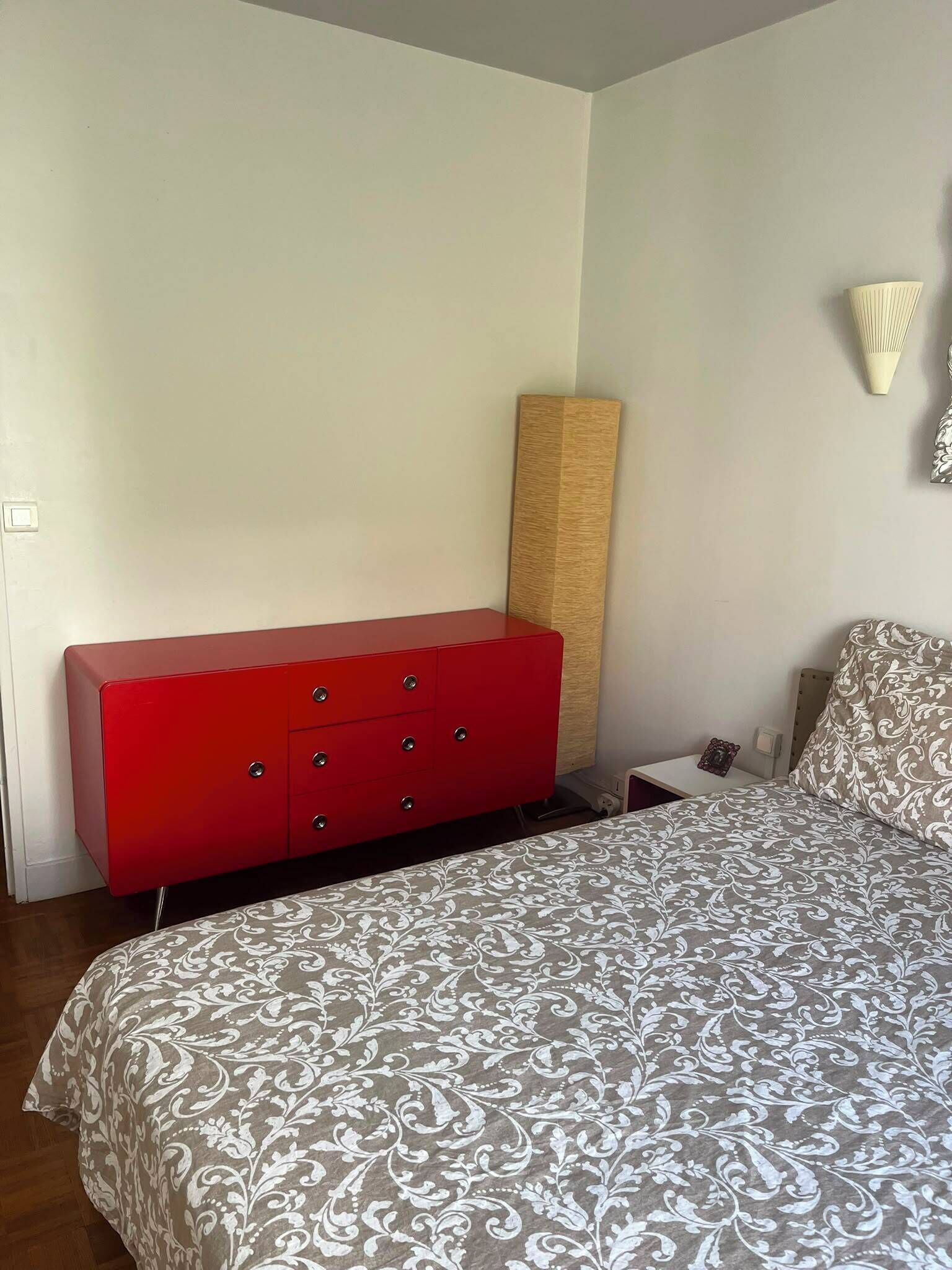 A bedroom with a bed, a red dresser, a nightstand, and a wall-mounted lamp.