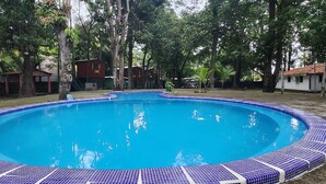 A swimming pool with a tiled edge, surrounded by trees and buildings. - Sunzal Bungalows (Tamanique)