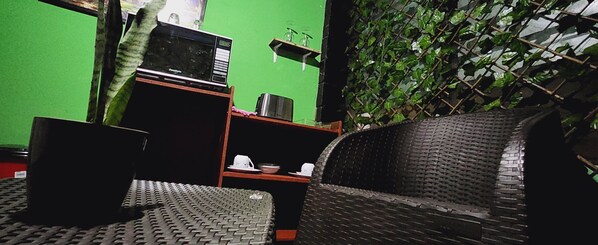 A room with a green wall, a microwave, a small shelf with glasses, and a wicker chair. - Sunzal Bungalows (Tamanique)