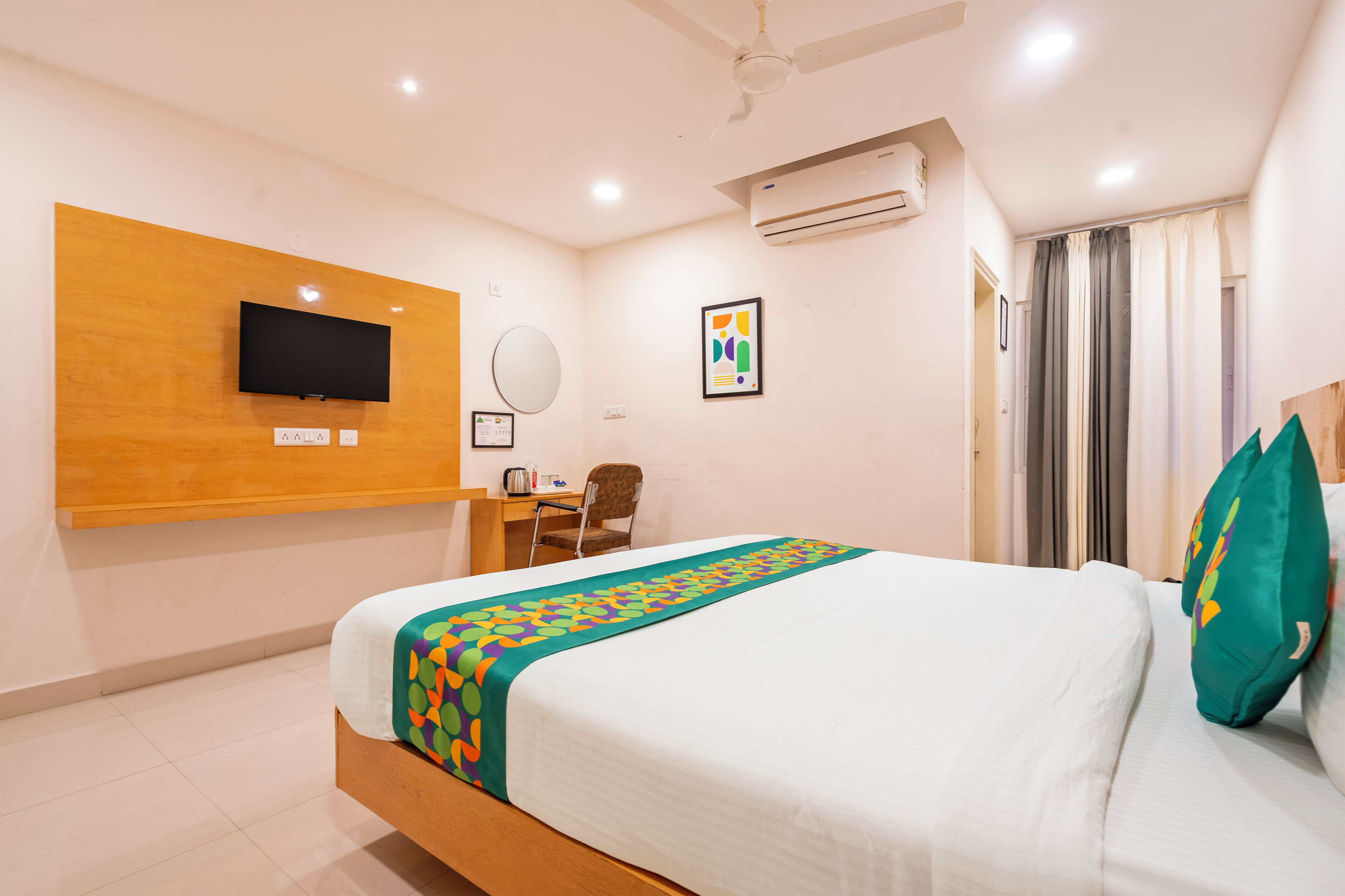 A hotel room with a bed, a desk, a chair, a TV, a mirror, and an air conditioning unit.
