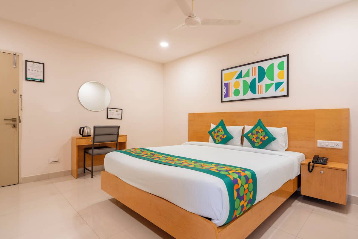 A hotel room with a bed, a desk, a chair, a bedside table with a phone, a wall-mounted mirror, and a ceiling fan.
