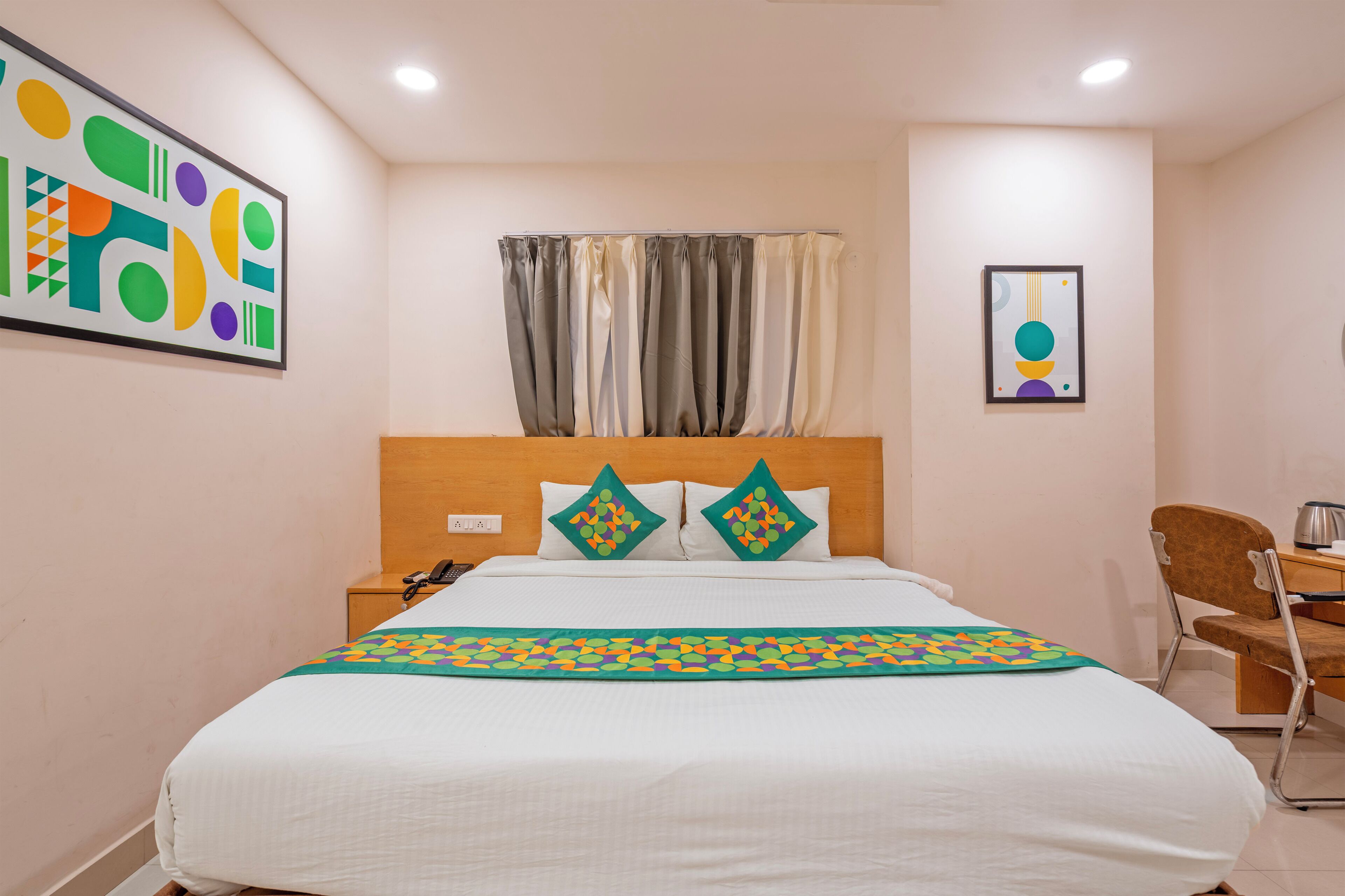 A hotel room with a bed, a desk, a chair, a nightstand, and a wall with artwork.