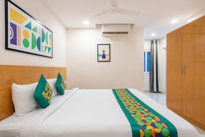 A hotel room with a bed, pillows, a ceiling fan, an air conditioning unit, a framed picture on the wall, and a wooden wardrobe.
