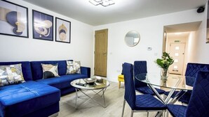 A modern living room with a blue sofa, a glass coffee table, and matching blue chairs. There are framed artworks on the wall and a round mirror above the coffee table. - COSY CRUISE CRIBS - MODERN HOME FROM HOME - CENTRAL LOCATION. (Stoke-on-Trent)