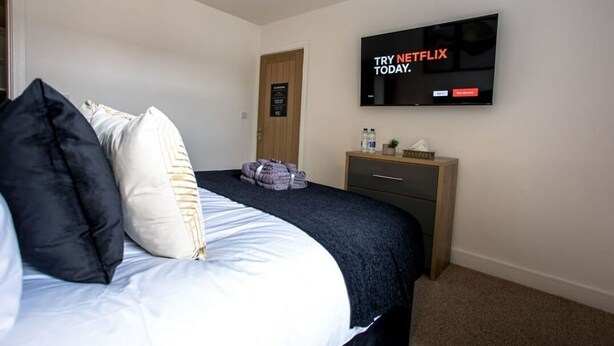 A hotel room with a bed, a TV mounted on the wall, a wooden nightstand, and a door. - COSY CRUISE CRIBS - MODERN HOME FROM HOME - CENTRAL LOCATION. (Stoke-on-Trent)