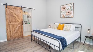 Quarto com estrutura de cama de metal, porta de madeira, mesa de cabeceira branca e uma obra de arte floral na parede.
