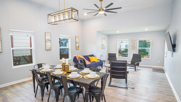 Uma sala de jantar com uma mesa de madeira escura posta para seis pessoas, rodeada por cadeiras pretas. Há um ventilador de teto acima e um lustre pendurado.