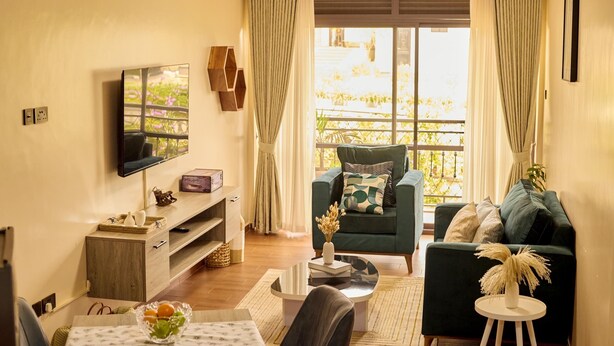 A modern living room with a flat-screen TV, a green sofa, and a dining area with a table set for two. - Sand Grains Residences 008 (Entebbe)