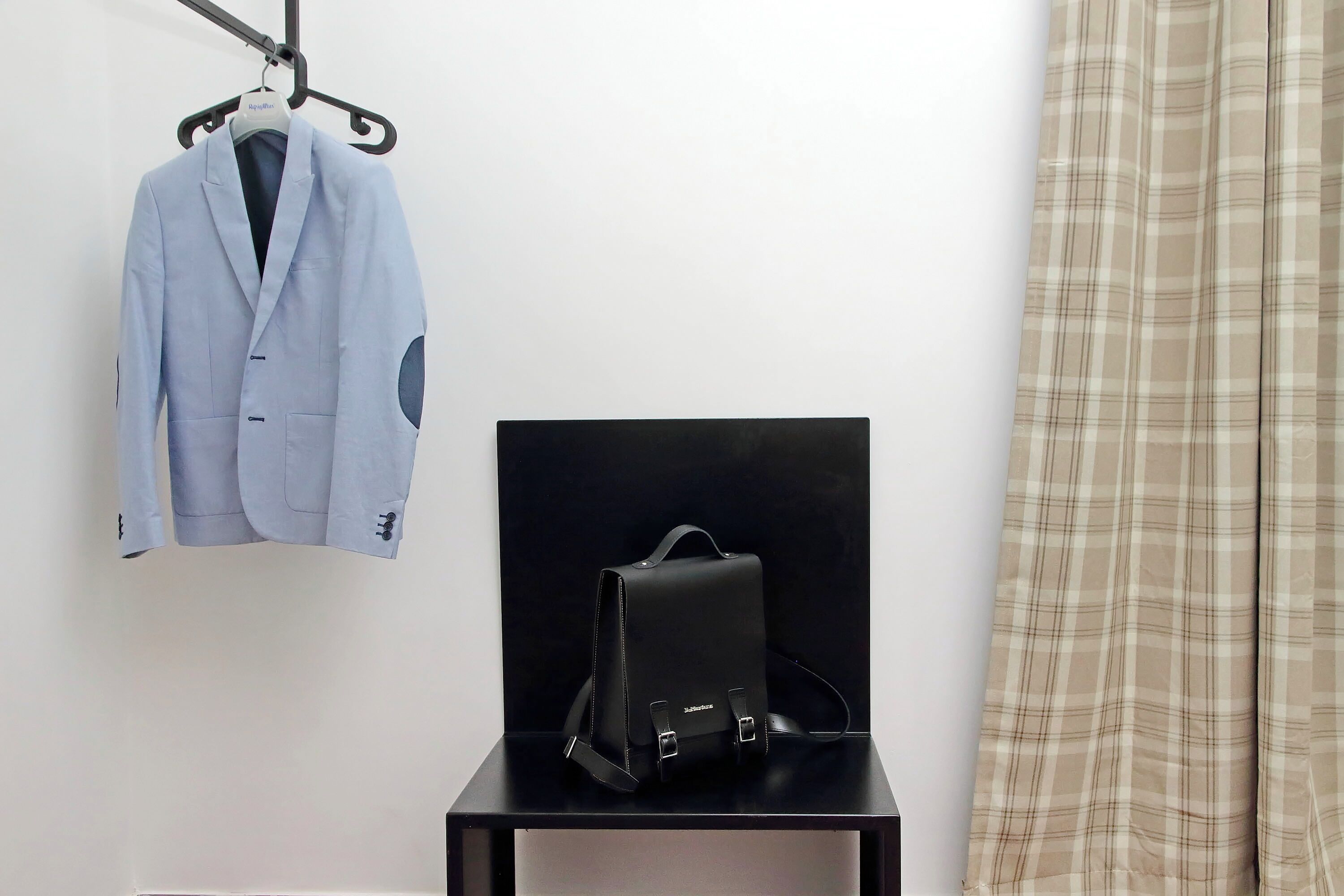Een kamer met een hangende lichtblauwe colbert, een zwarte lederen tas op een zwart standaard en een ruitendecor gordijn.
