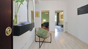 A hallway with a black console table, a green chair, and a vase with sunflowers. There is a door with a brass handle, a blackboard with a welcome sign, and a framed picture on the wall. - TM Apartment (Rome)