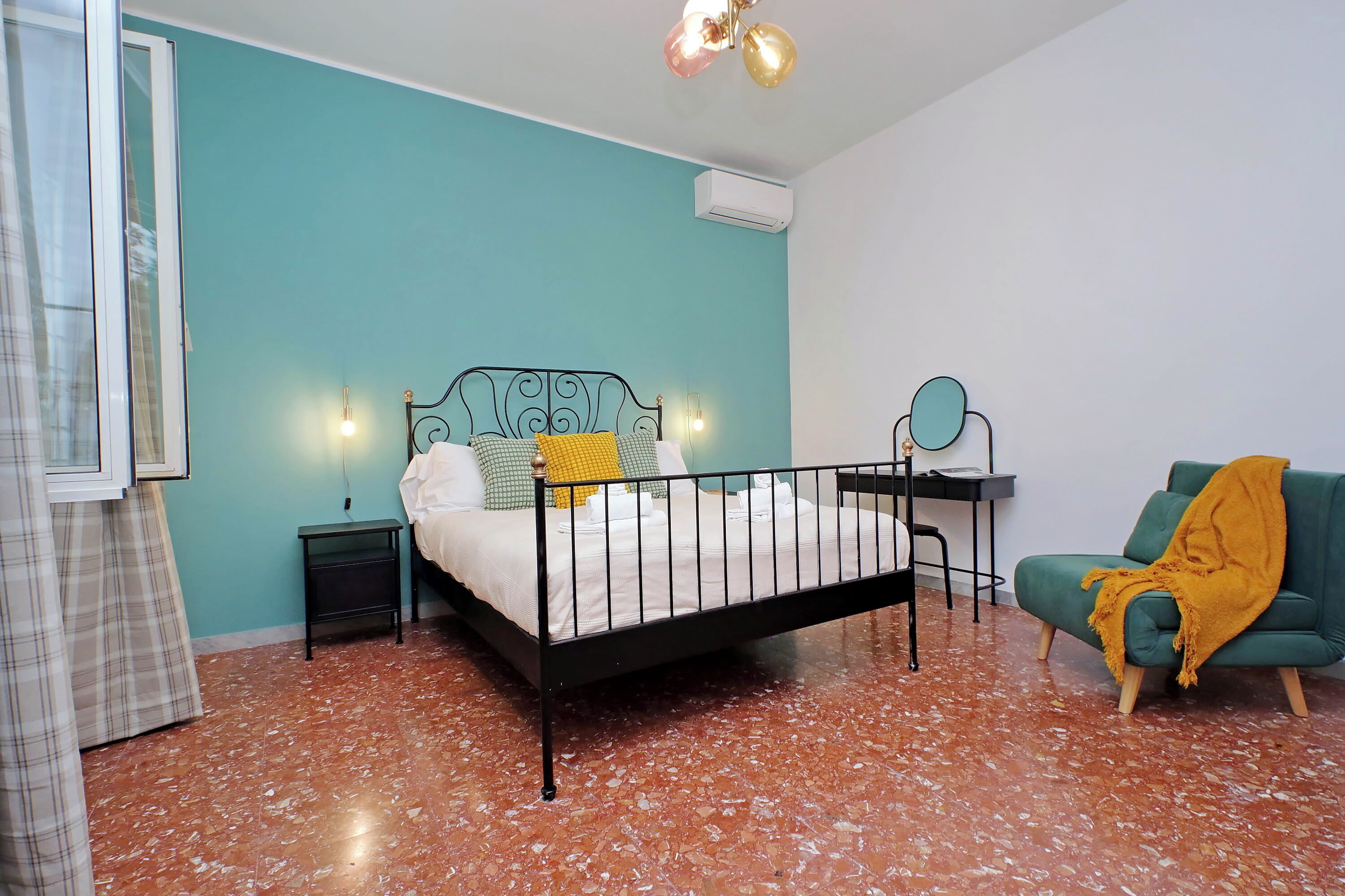 A bedroom with a bed, a green armchair, a small table with a mirror, and a window with curtains.
