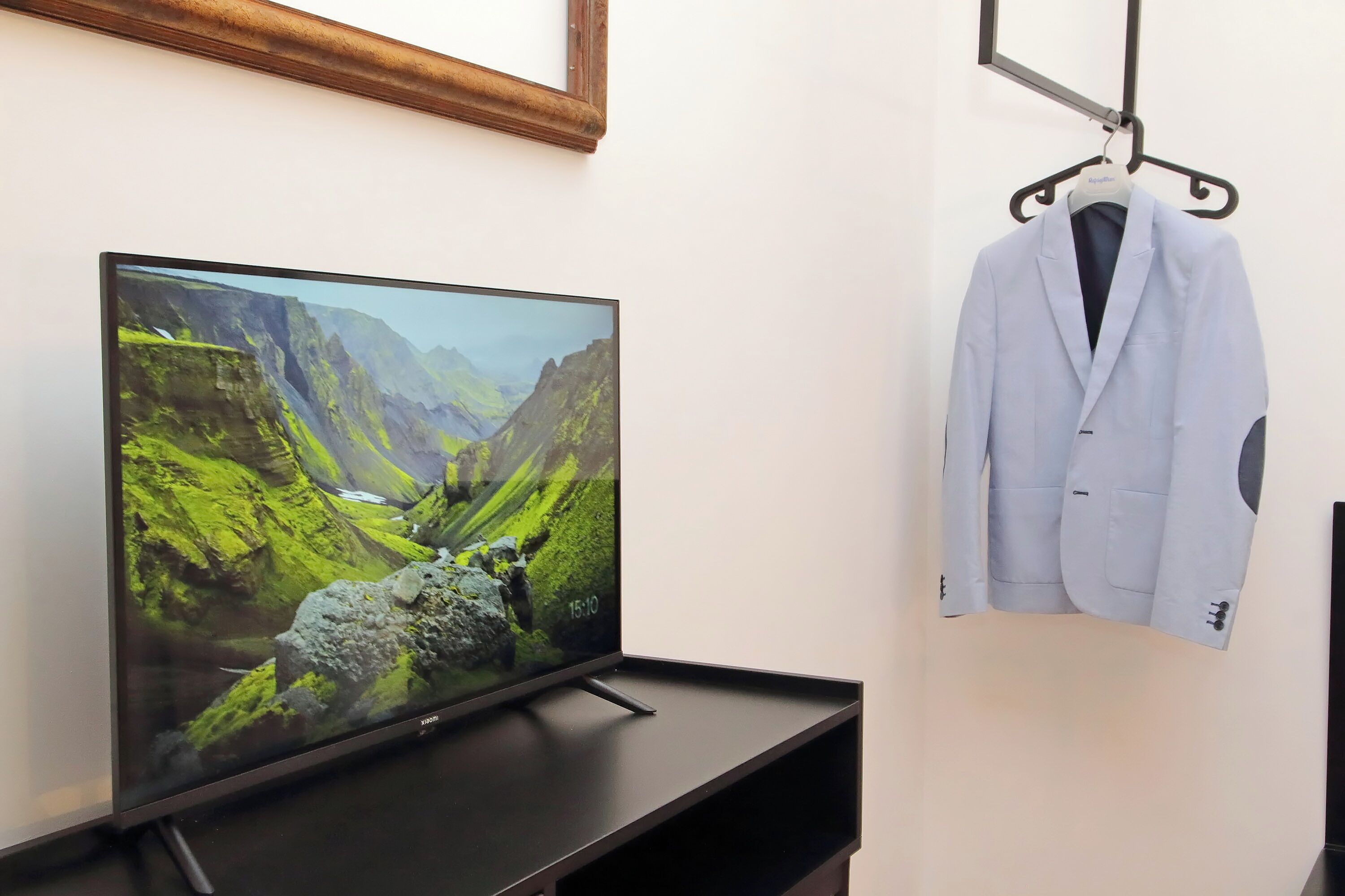 Ein Flachbildfernseher auf einem schwarzen Ständer, ein weißer Mantel an einem schwarzen Kleiderbügel und ein Bild mit Holzrahmen über dem Fernseher.