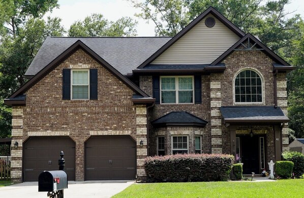 A two-story house with a brick facade, two garage doors, and a front entrance with a small porch. - Spacious 5-bedroom house in peaceful Midway with WiFi, AC Close to Savannah GA (Midway)