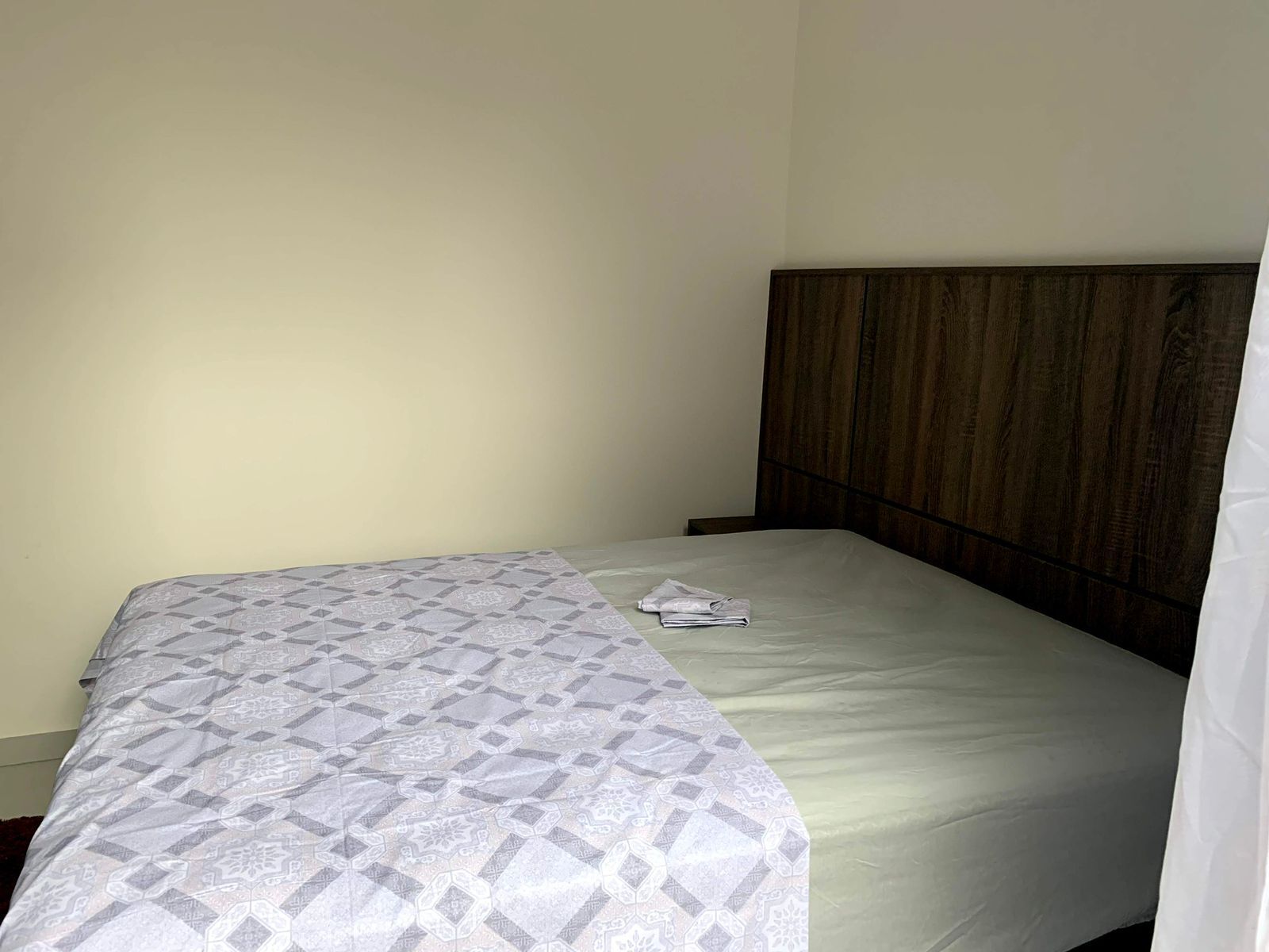 A single bed with a patterned bedspread, a wooden headboard, and a plain wall.