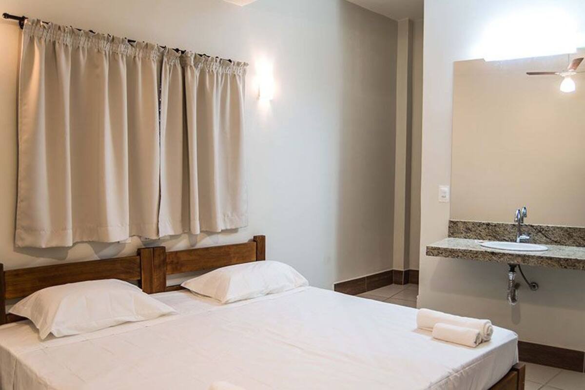 A hotel room with a bed, a wooden headboard, a bathroom with a sink and mirror, and beige curtains.