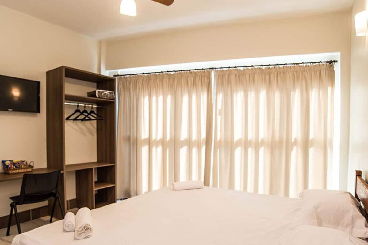 A hotel room with a bed, a TV mounted on the wall, a desk with a chair, a wardrobe, and a large window with curtains.