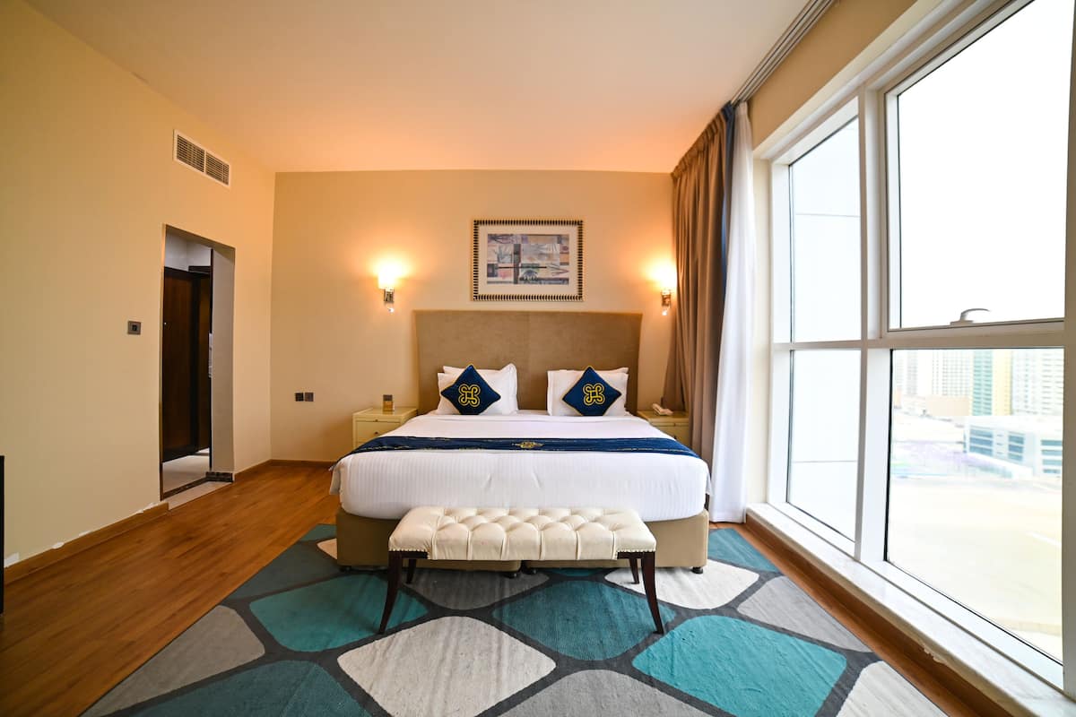 A hotel room with a large bed, a bench, a view of the city, and a painting on the wall.