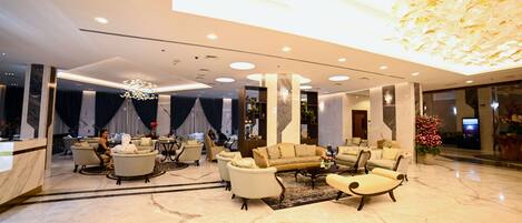A spacious lobby with marble flooring, a central seating area with sofas and armchairs, a reception desk, and a decorative ceiling feature.