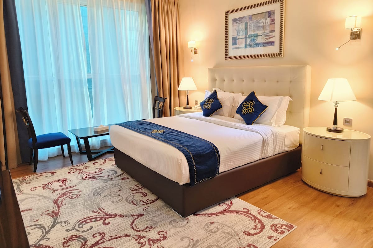 A hotel room with a large bed, two bedside lamps, a chair, a small table, and a view of the outdoors through sheer curtains.
