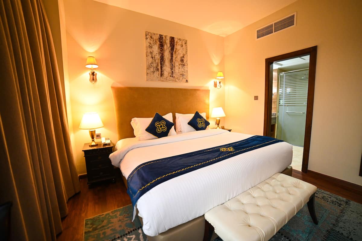 A hotel room with a large bed, two bedside lamps, a headboard, a bench, a nightstand, and a painting on the wall.