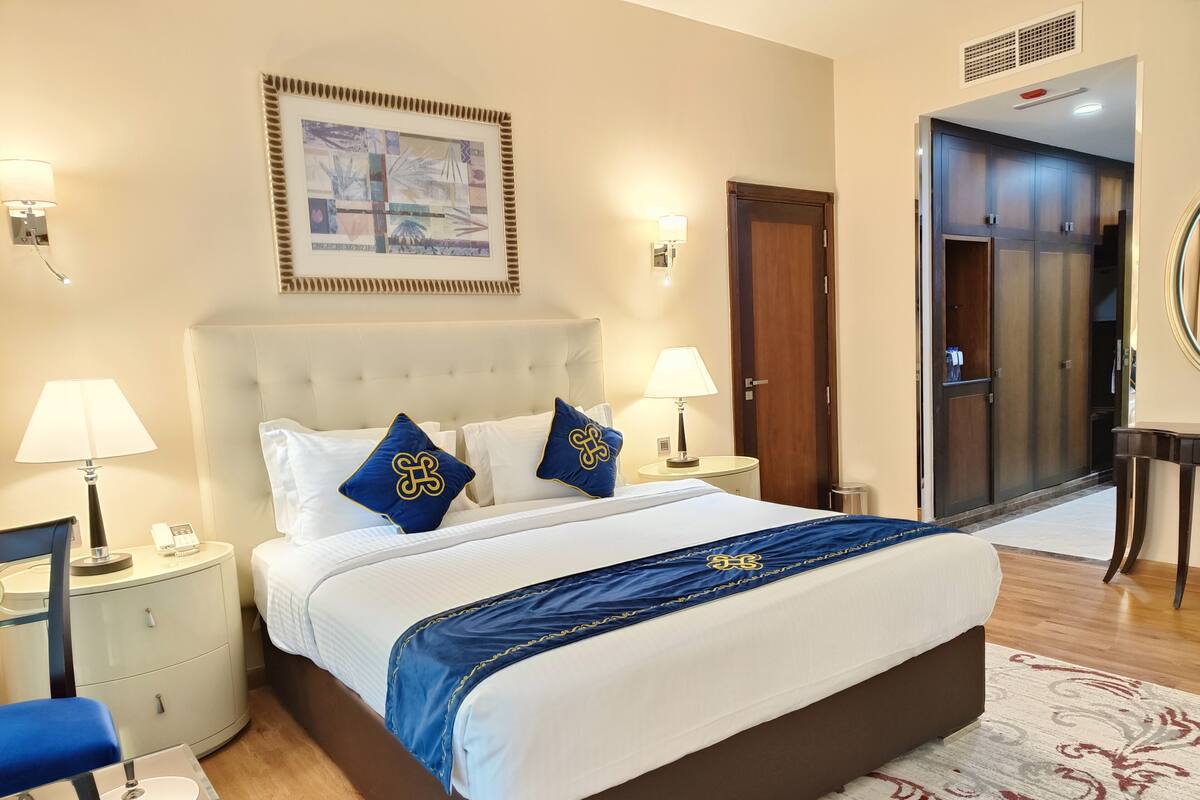 A hotel room with a large bed, two bedside lamps, a framed artwork above the bed, a wardrobe, and a small table with a lamp.