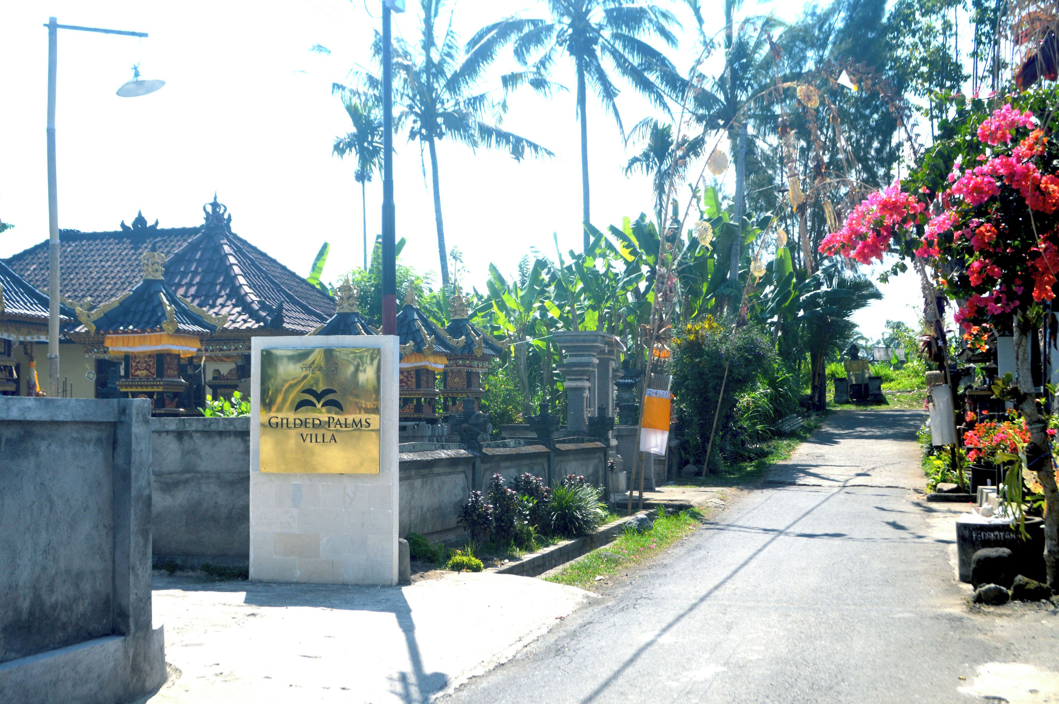 Señal para Gilded Palms Villa, un edificio tradicional con techo de tejas, rodeado de exuberante follaje y palmeras.