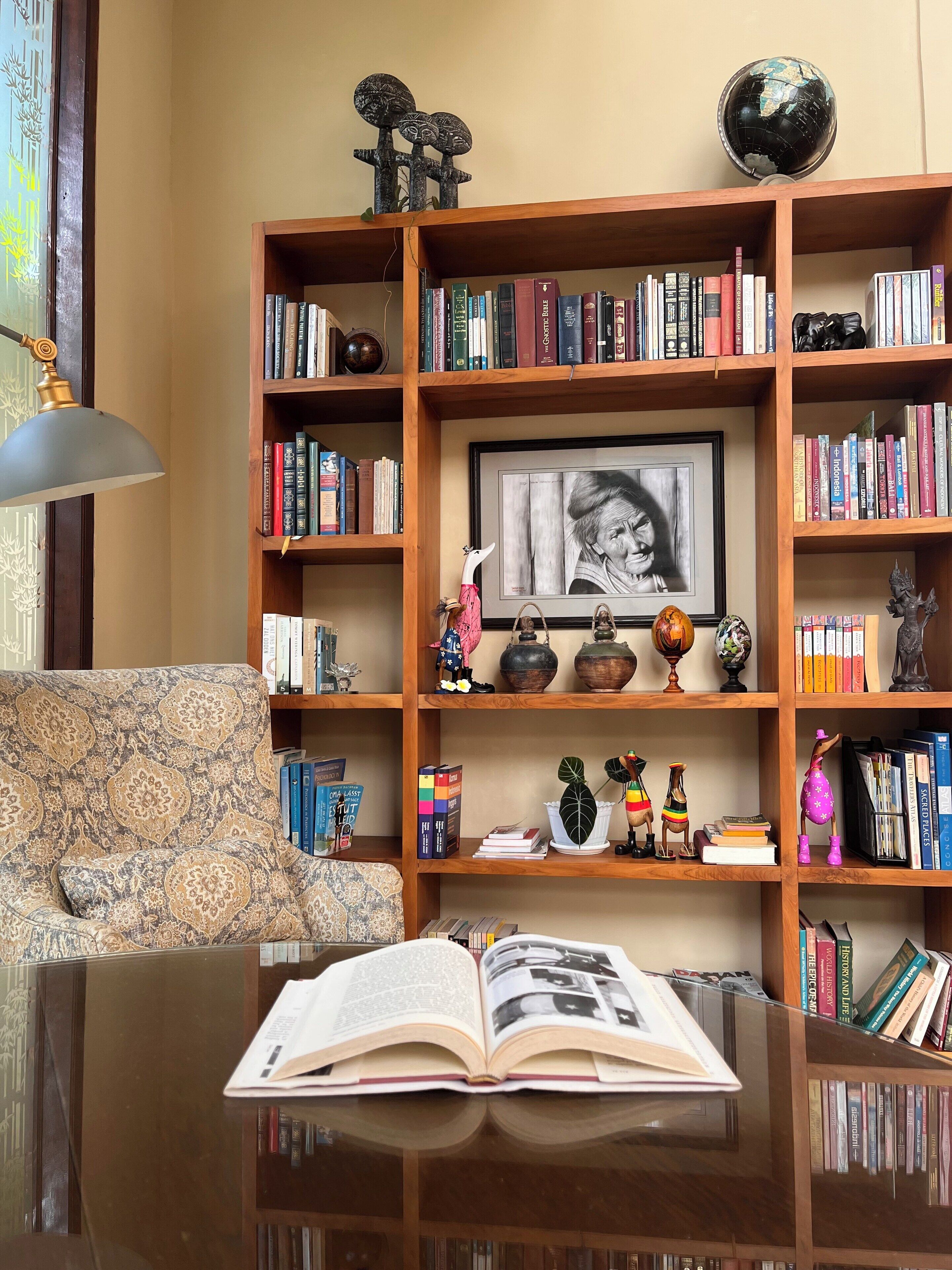 Una habitación con una estantería de madera llena de libros, un retrato enmarcado, objetos decorativos y una mesa con un libro abierto.