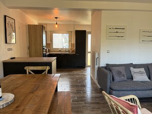 A modern kitchen with a large window, a dining area with a wooden table and chairs, and a grey sofa in a living room. - Beg Meil dunes house (Fouesnant)
