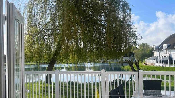 A view from a balcony with a white railing, overlooking a calm lake, a willow tree, and a modern house across the water. - Lake’s End Lodge.  (South Cerney)