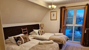 A bedroom with two beds, a wooden headboard, a bedside table with a lamp, a framed picture on the wall, and a view of trees through a window with curtains. - Lake’s End Lodge.  (South Cerney)