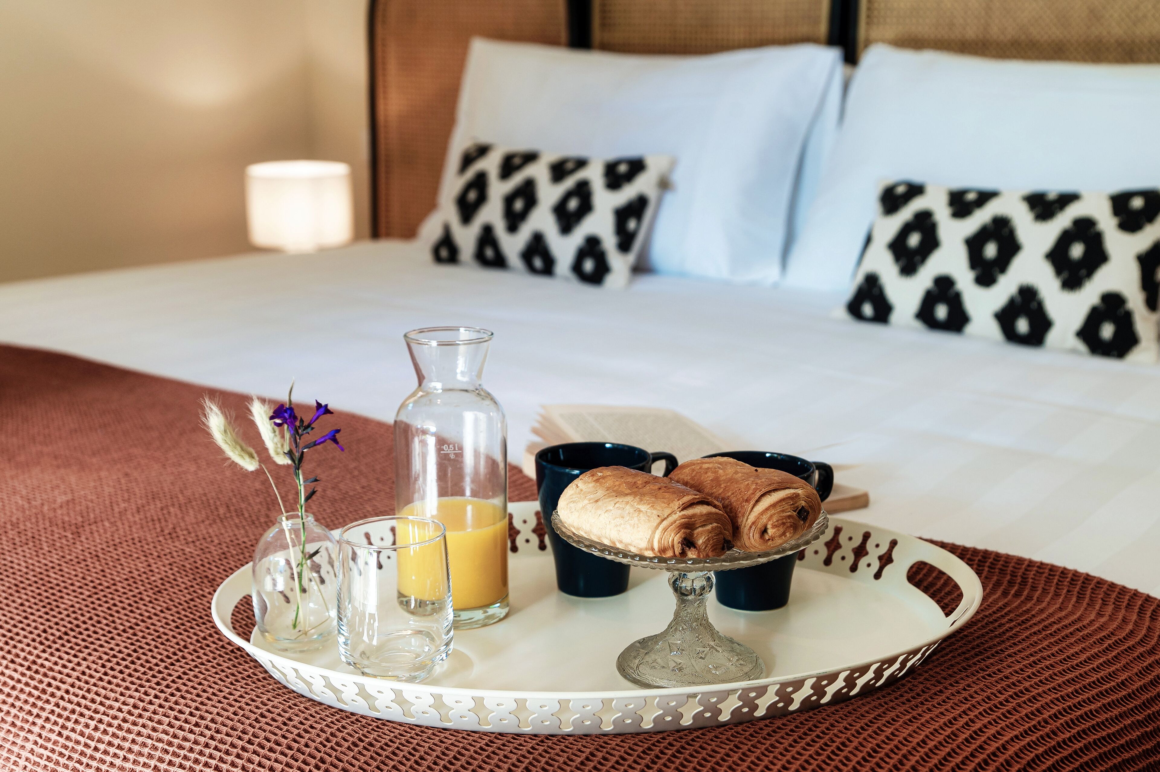 A neatly made breakfast tray with a bottle of orange juice, two cups, a glass, and a rolled-up pastry on a bed with patterned pillows and a bedside lamp.