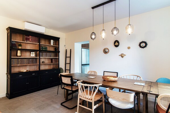 Un coin repas avec une table en bois, des chaises en osier et un grand meuble noir rempli de livres.