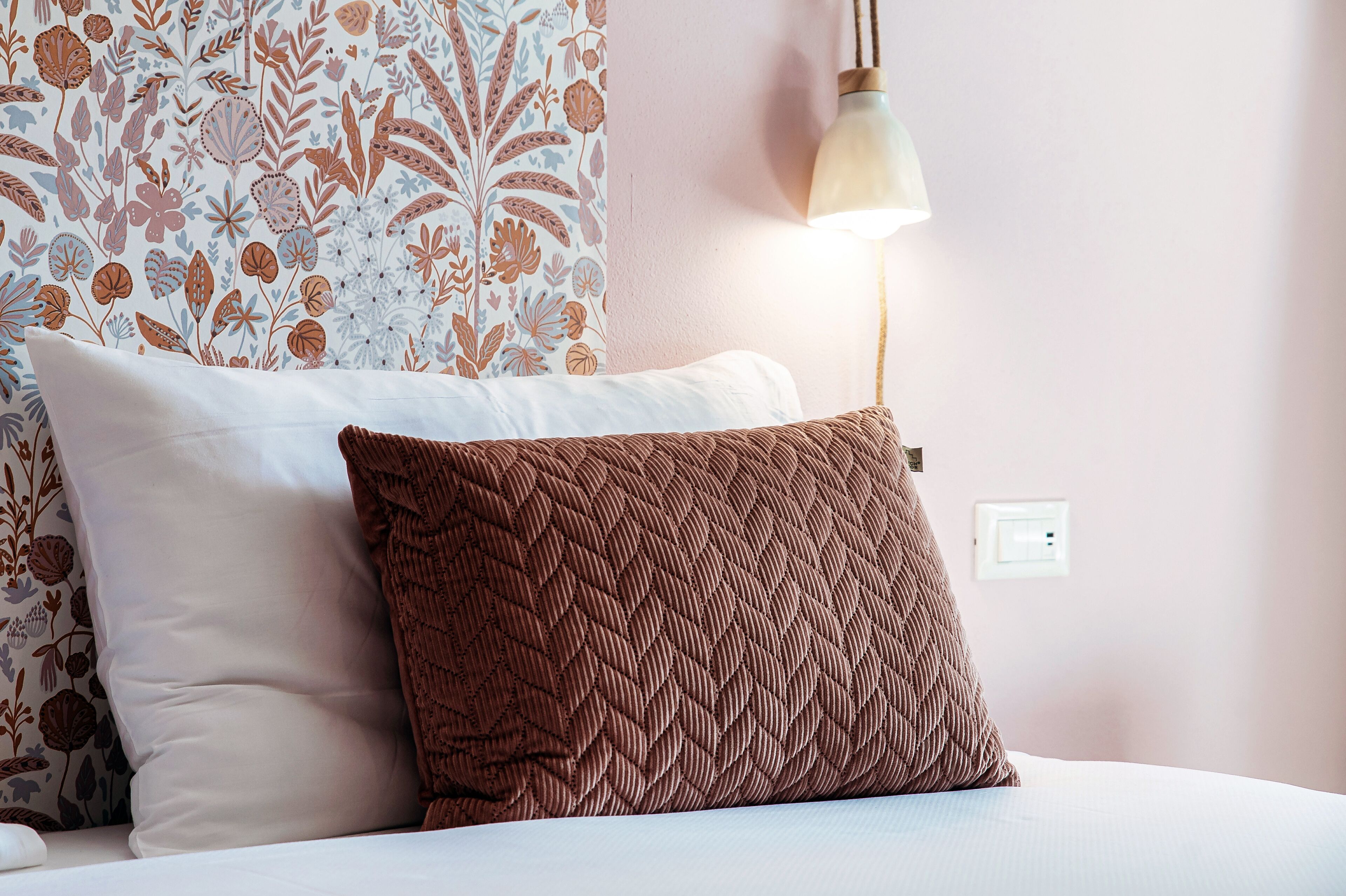 A bed with white and brown pillows, a floral-patterned headboard, and a wall-mounted light fixture.
