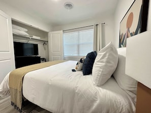 A hotel room with a bed, a television, a window with blinds, and a painting on the wall.