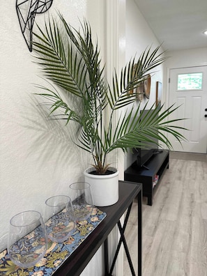 Un pasillo con una planta en maceta, una mesa de vidrio con copas de vino y un banco negro.