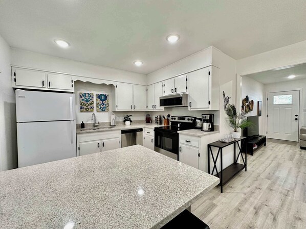 A modern kitchen with white cabinets, granite countertops, a refrigerator, stove, microwave, and a dining area with a table and chairs.