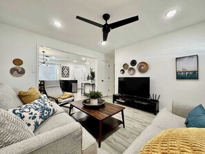 A modern living room with a sofa, coffee table, TV, ceiling fan, and decorative wall art.