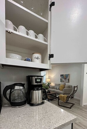 A kitchen with a coffee maker, a cabinet with coffee mugs, and a sofa in the background.