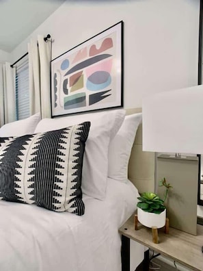 A neatly arranged hotel room with a bed, pillows, a bedside table with a lamp, a framed abstract artwork, and a potted plant.