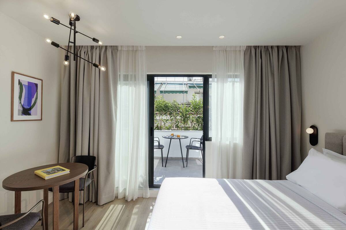Ein Hotelzimmer mit einem Bett, einem Schreibtisch mit Stuhl, einem Tisch für zwei Personen und Blick durch eine Glastür nach draußen.