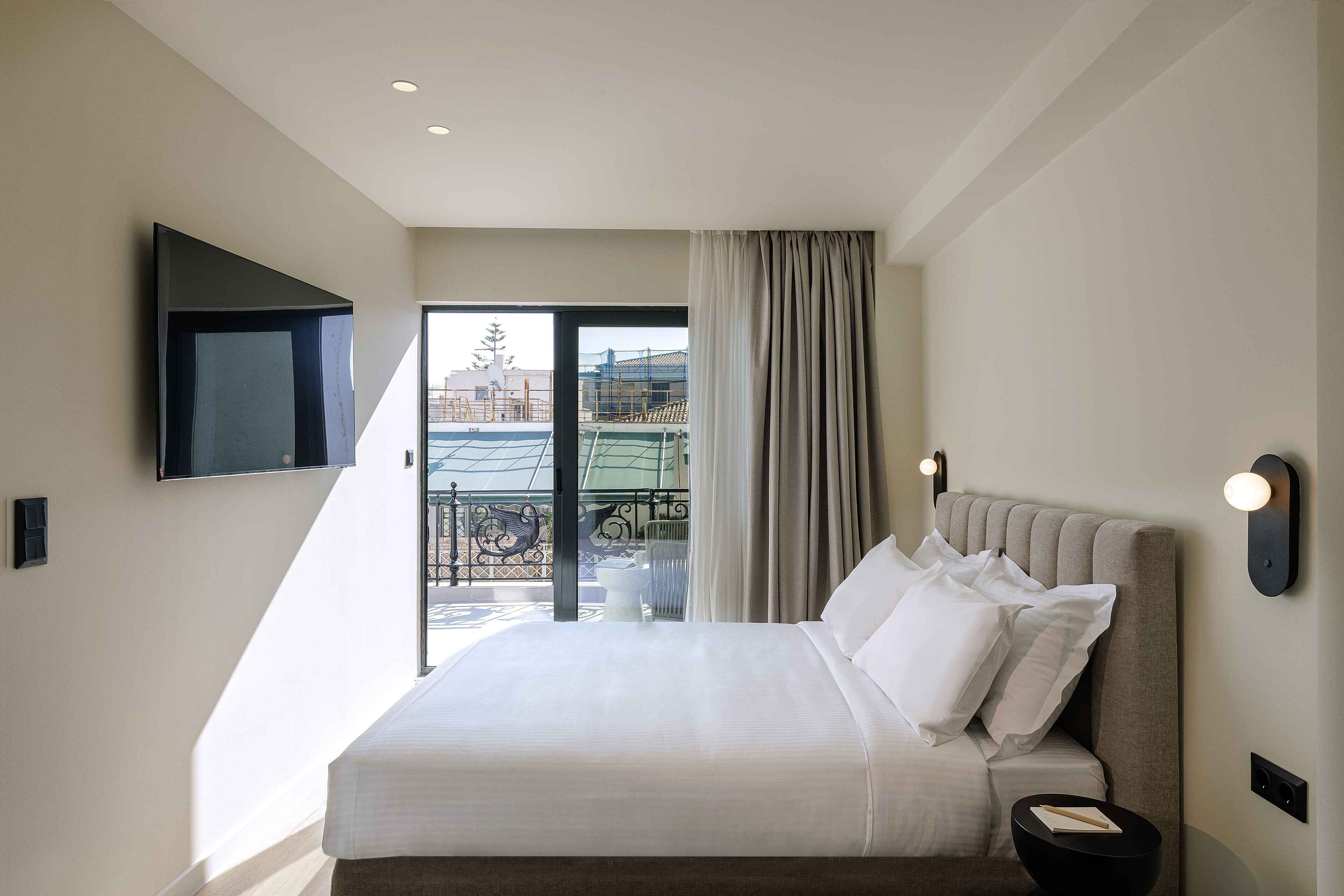 A modern hotel room with a large bed, a flat-screen TV mounted on the wall, and a balcony with a view of the city.