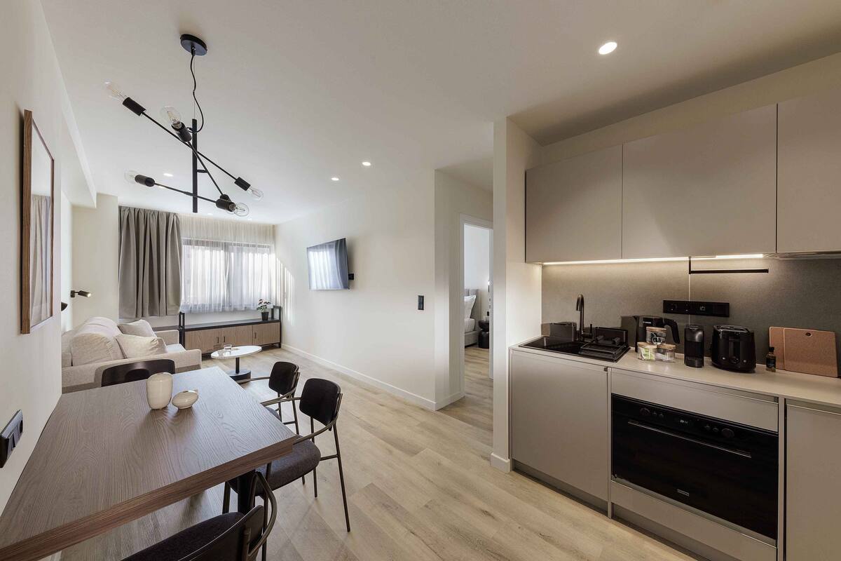 Eine moderne Küche mit Backofen, eine Essecke mit Holztisch und Stühlen sowie ein Wohnbereich mit Sofa und Fernseher.