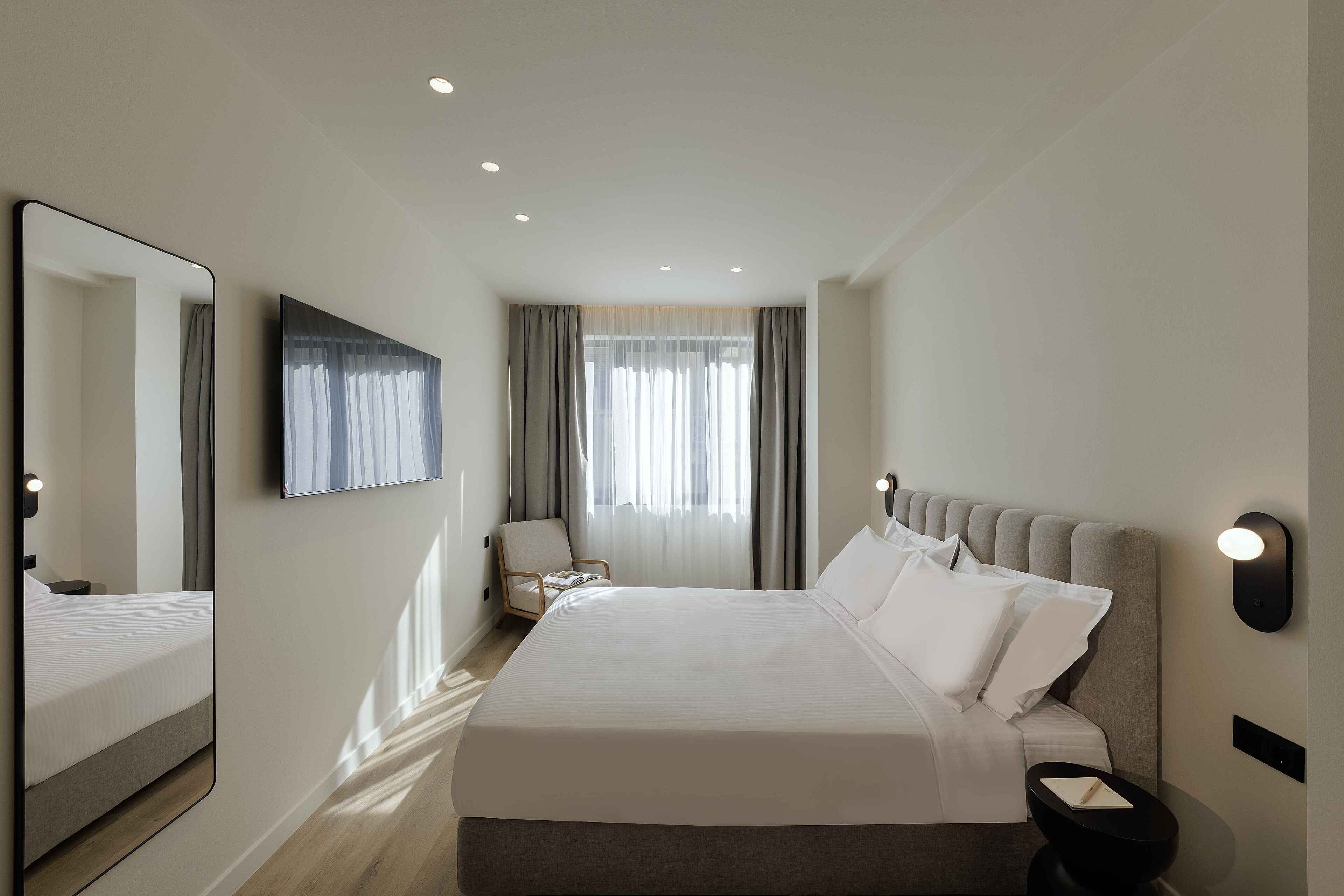 A modern hotel room with a large bed, a sitting area with a chair, a flat-screen TV, and a large mirror.