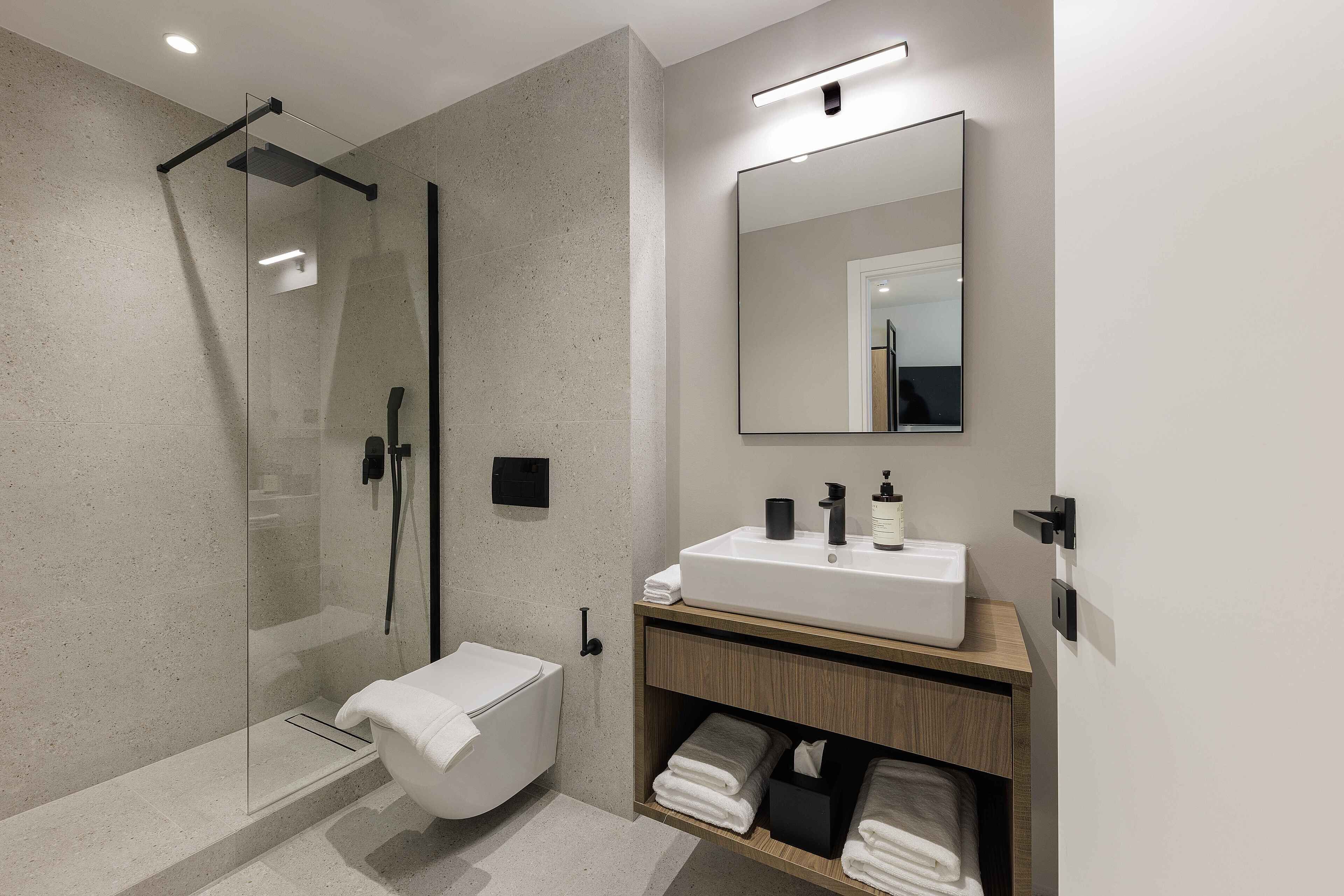 Un baño moderno con ducha de vidrio, lavamanos blanco sobre tocador de madera y un espejo grande.